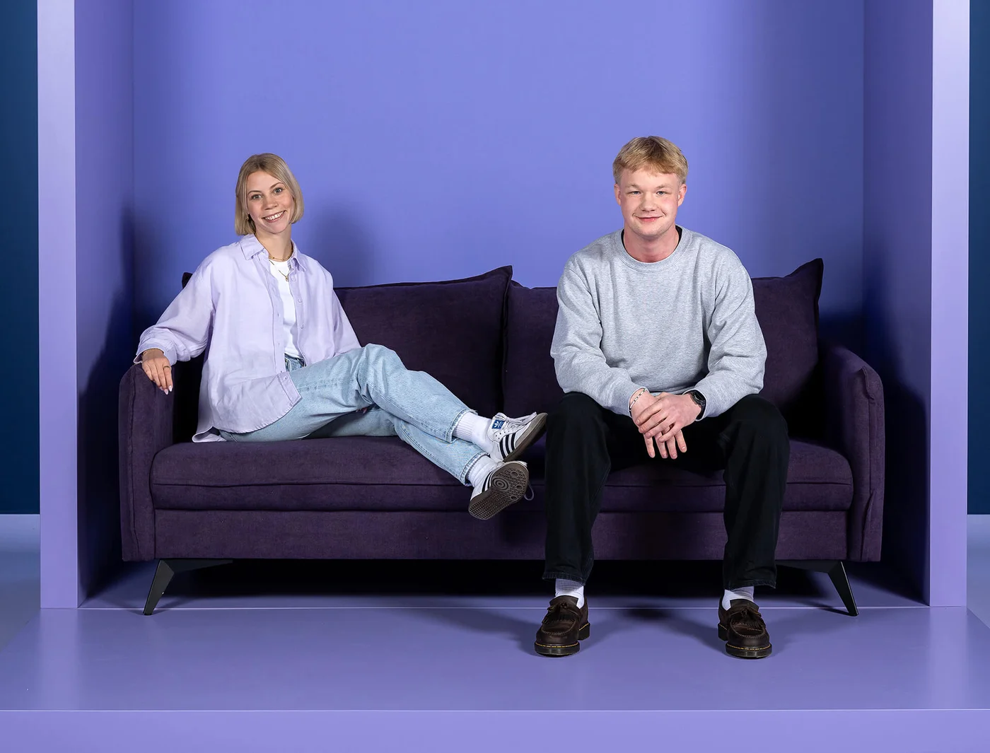 Zwei Personen sitzen auf einem lila Sofa vor einer lila Wand. Die Frau trägt eine helle Bluse und Jeans, der Mann ein graues Sweatshirt und dunkle Hosen. Beide lächeln in die Kamera.