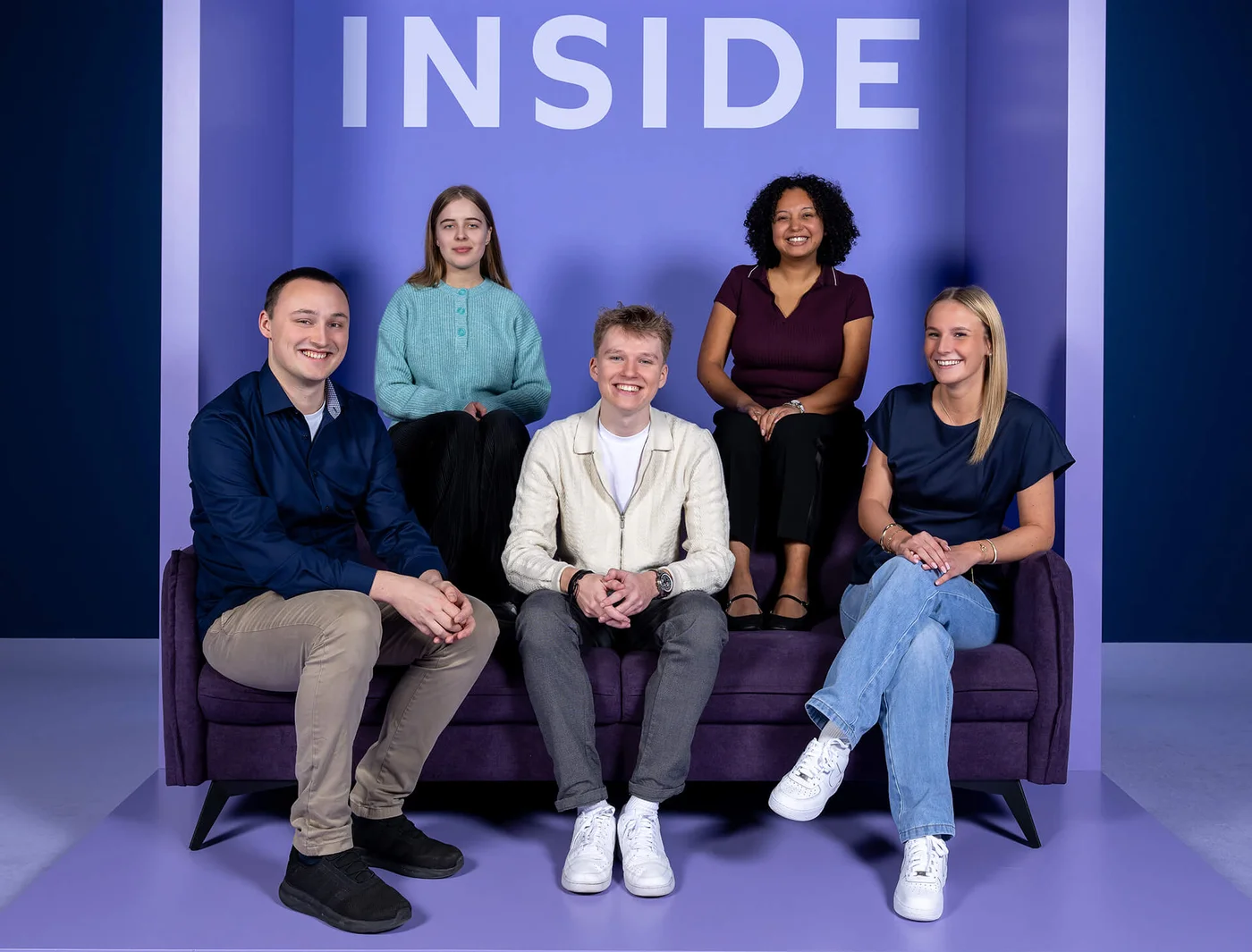 Fünf Personen sitzen auf einem lila Sofa vor einem lila Hintergrund mit dem Wort "INSIDE" darüber. Alle lächeln, und die Stimmung wirkt fröhlich und entspannt.