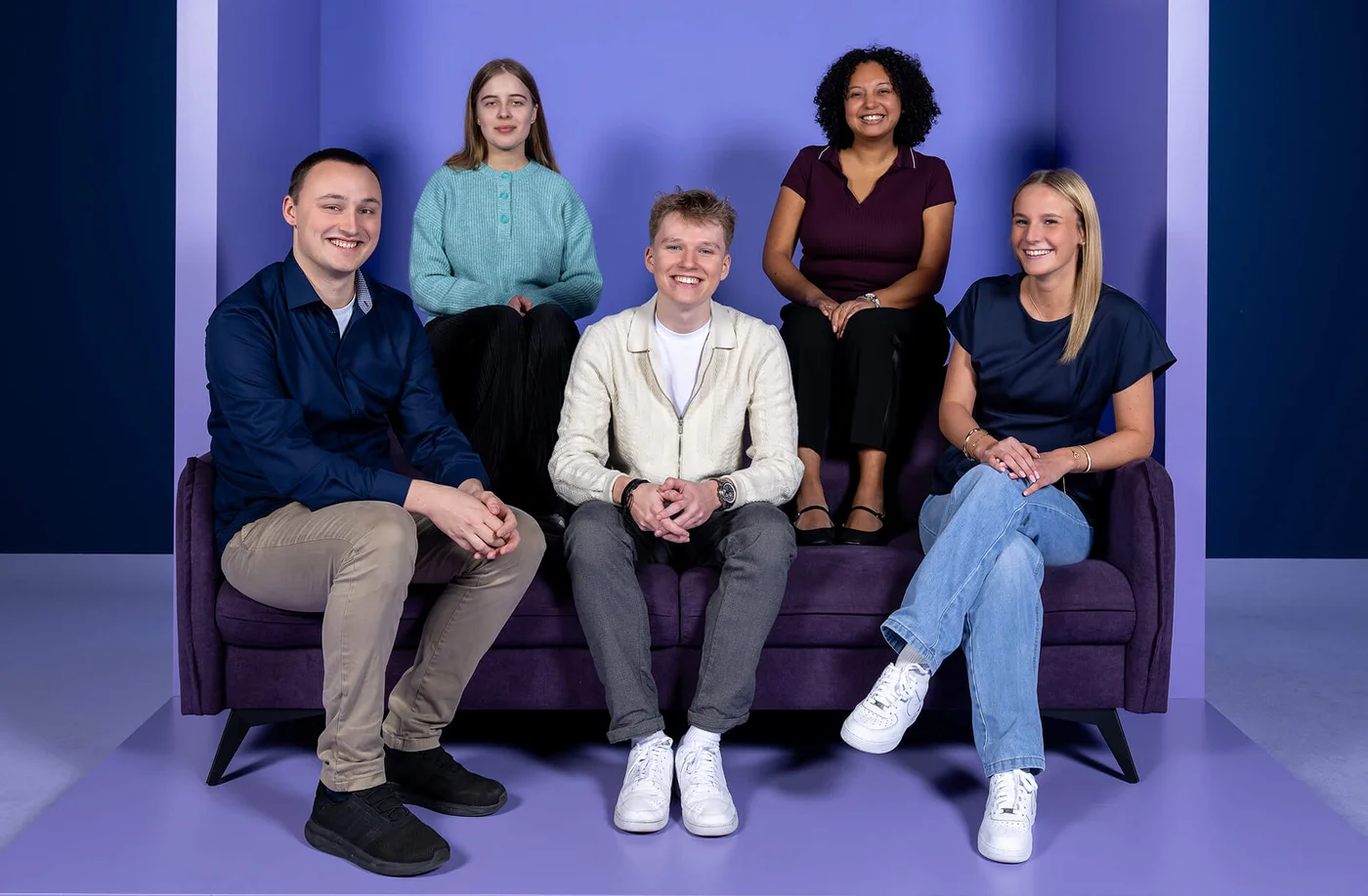 Fünf Personen sitzen auf einem lila Sofa vor einem lila Hintergrund mit dem Wort "INSIDE" darüber. Alle lächeln, und die Stimmung wirkt fröhlich und entspannt.