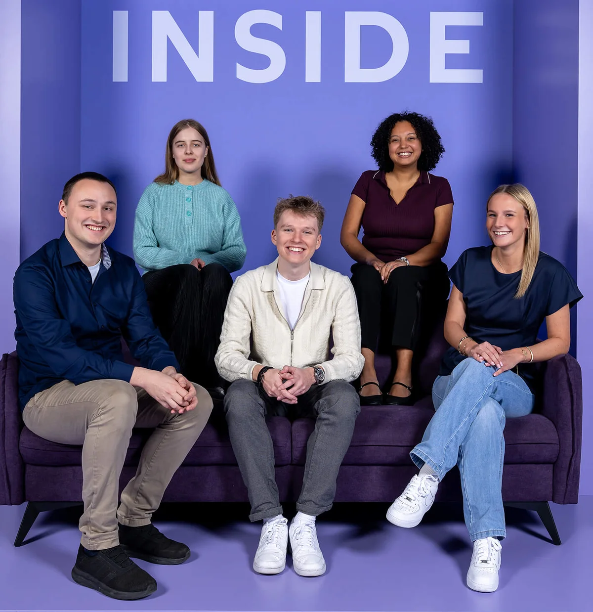 Fünf Personen sitzen auf einem lila Sofa vor einem lila Hintergrund mit dem Wort "INSIDE" darüber. Alle lächeln, und die Stimmung wirkt fröhlich und entspannt.