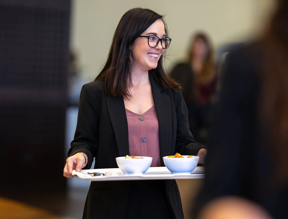 Eine lächelnde Frau in einem schwarzen Blazer trägt ein Tablett mit zwei Schüsseln vor sich. Sie steht in einem Innenraum und scheint in Bewegung zu sein.