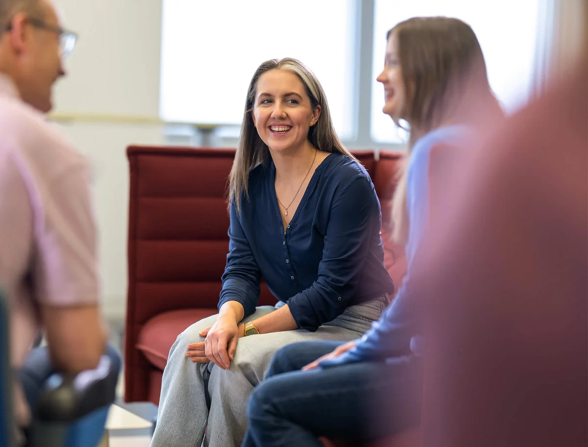 Eine Gruppe von Menschen sitzt zusammen und unterhält sich in einer entspannten, freundlichen Atmosphäre. Eine Frau in der Mitte lächelt und wirkt aufmerksam. Es scheint ein informelles, geselliges Treffen oder Gespräch zu sein.