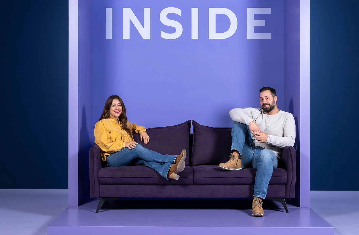 Zwei Personen sitzen entspannt auf einem lila Sofa in einem minimalistischen, lilafarbenen Raum. Die Frau trägt eine gelbe Bluse, der Mann ein weißes Oberteil. Über ihnen ist das Wort "INSIDE" zu lesen.