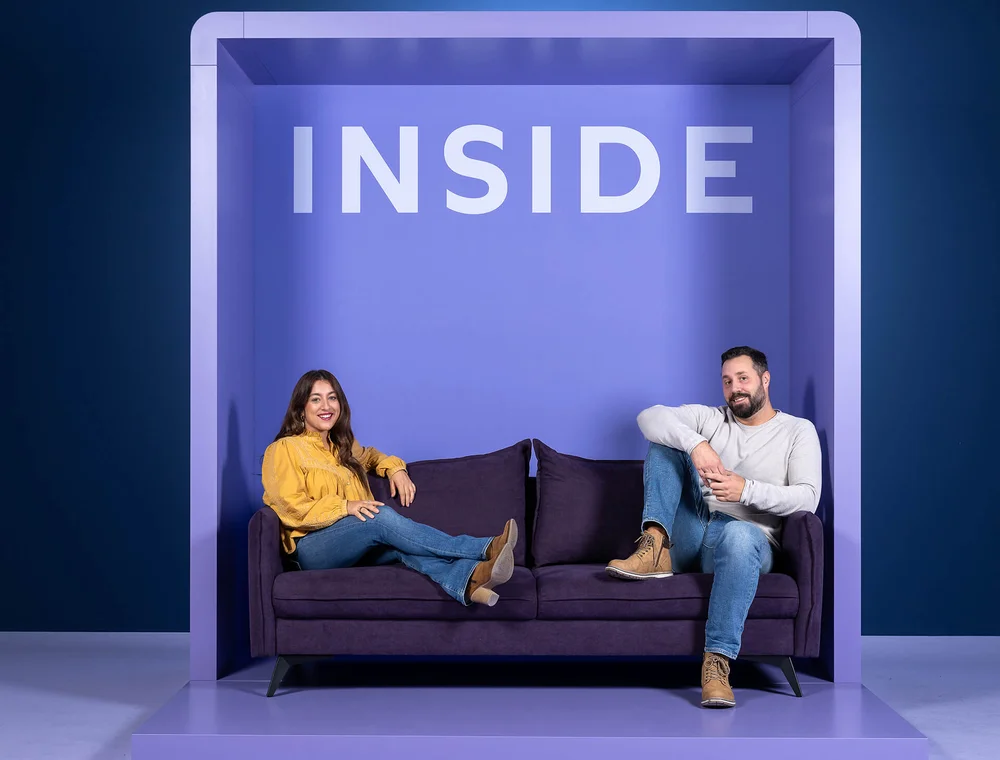 Zwei Personen sitzen entspannt auf einem lila Sofa in einem minimalistischen, lilafarbenen Raum. Die Frau trägt eine gelbe Bluse, der Mann ein weißes Oberteil. Über ihnen ist das Wort "INSIDE" zu lesen.
