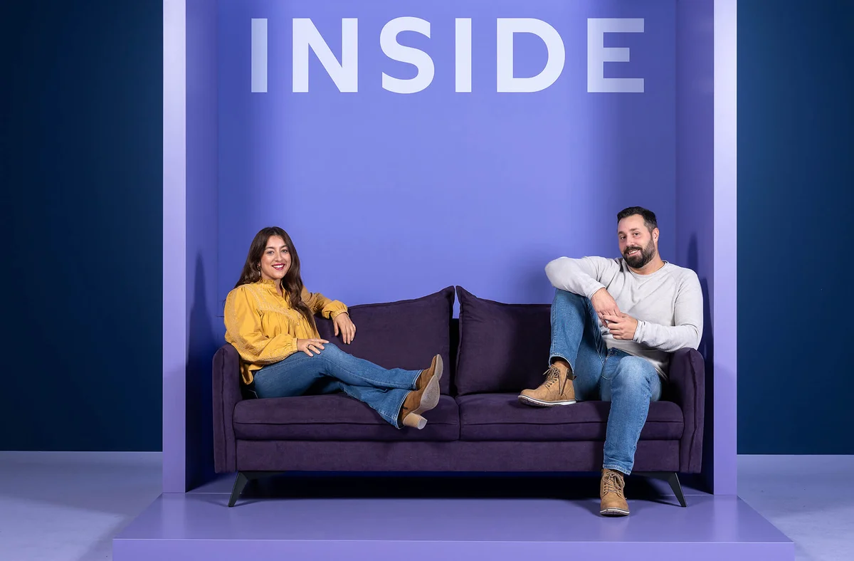 Zwei Personen sitzen entspannt auf einem lila Sofa in einem minimalistischen, lilafarbenen Raum. Die Frau trägt eine gelbe Bluse, der Mann ein weißes Oberteil. Über ihnen ist das Wort "INSIDE" zu lesen.