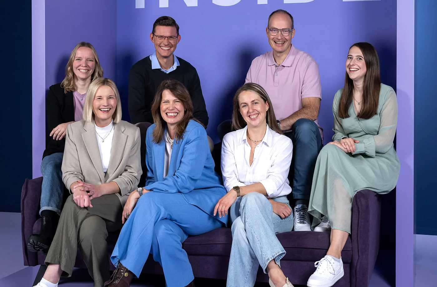 Eine Gruppe von sieben Personen sitzt auf einem Sofa vor einer lila Wand mit dem Wort "INSIDE". Alle lächeln in die Kamera.