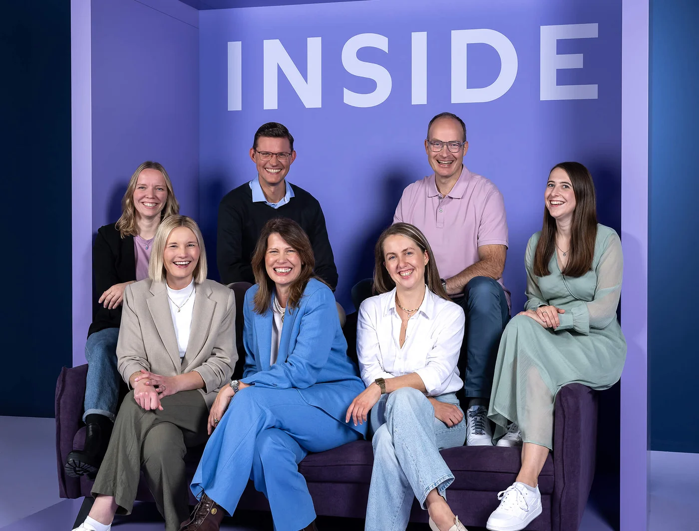 Eine Gruppe von sieben Personen sitzt auf einem Sofa vor einer lila Wand mit dem Wort "INSIDE". Alle lächeln in die Kamera.