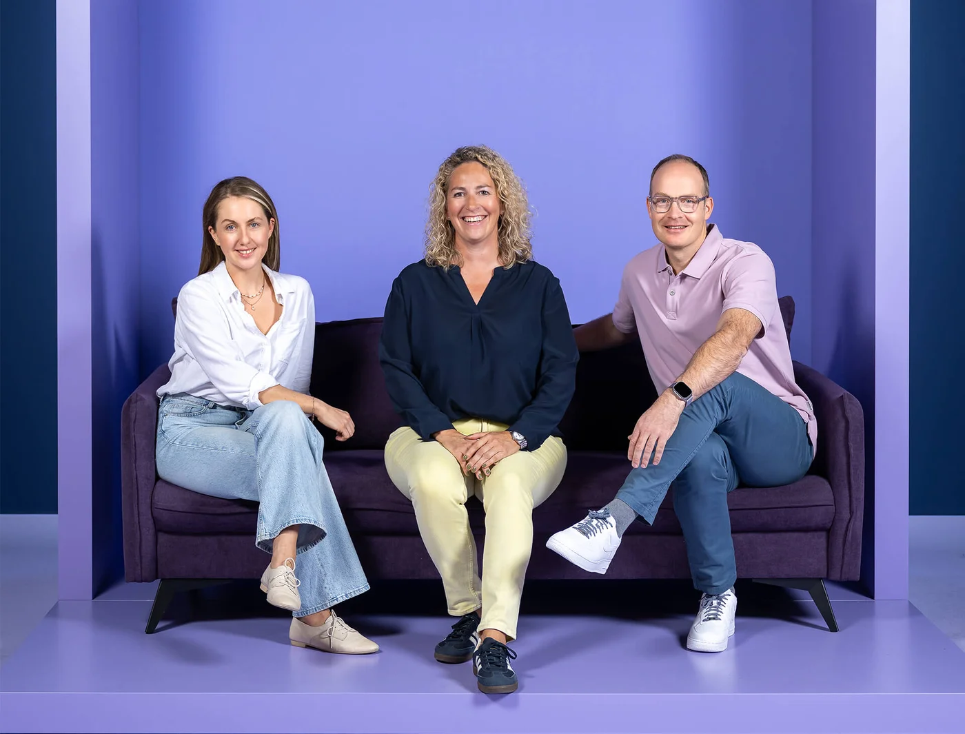 Drei Personen sitzen auf einem lila Sofa vor einem ebenfalls lila Hintergrund. Zwei Frauen und ein Mann lächeln in die Kamera. Die Frau links trägt ein weißes Hemd und Jeans, die Frau in der Mitte trägt eine dunkle Bluse, und der Mann trägt ein rosa Polohemd.