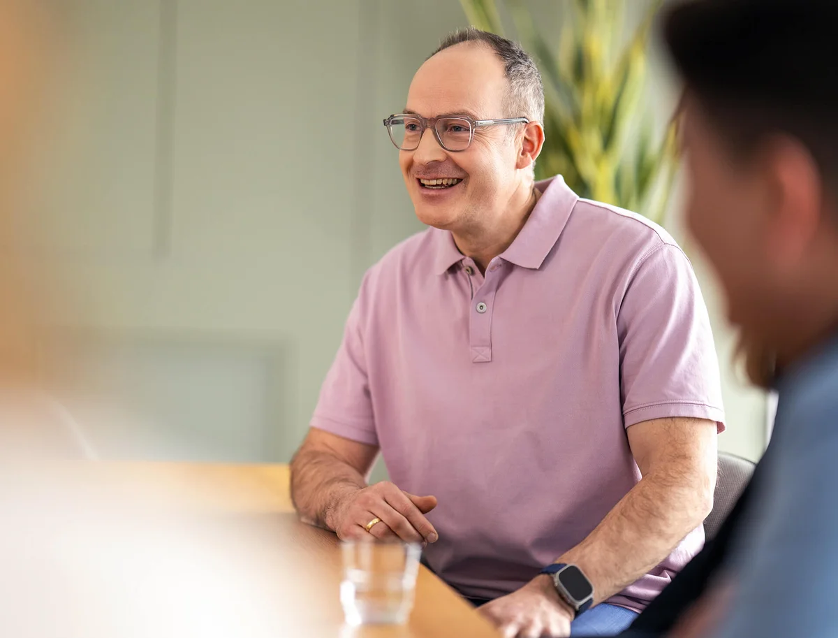 Ein lächelnder Mann mit Brille und rosa Poloshirt sitzt an einem Tisch. Er scheint in ein Gespräch vertieft zu sein. Im Vordergrund sind verschwommene Umrisse anderer Personen sichtbar, die an der Besprechung teilnehmen.
