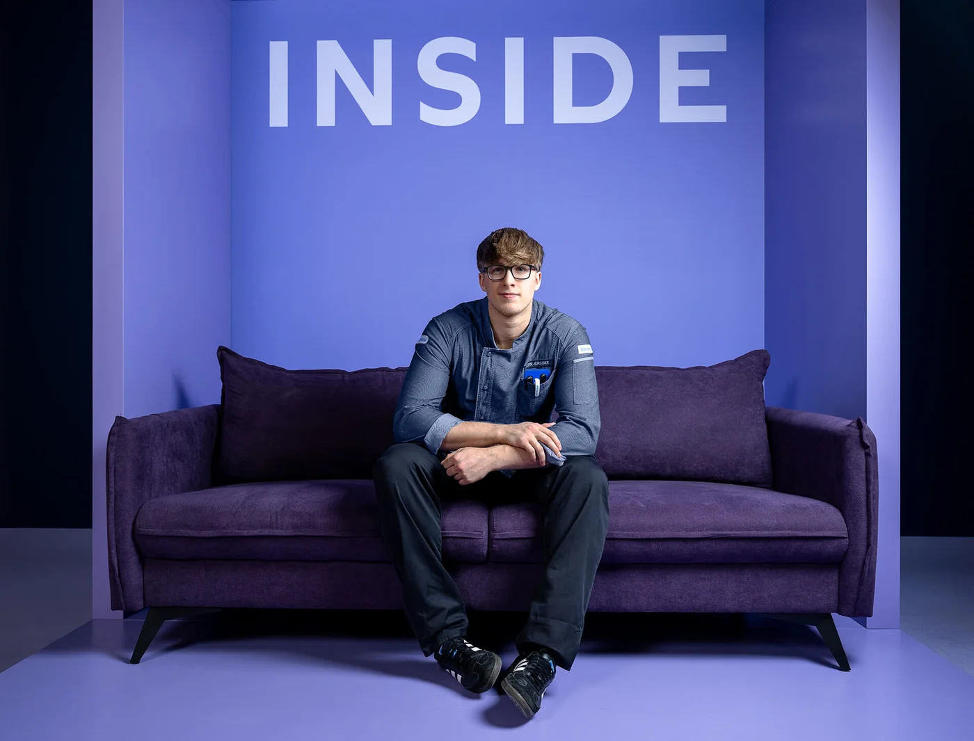 Eine Person sitzt auf einem lila Sofa vor einer lila Wand mit dem Wort "INSIDE" in großen Buchstaben darüber. Die Umgebung ist minimalistisch und in verschiedenen Lilatönen gehalten.