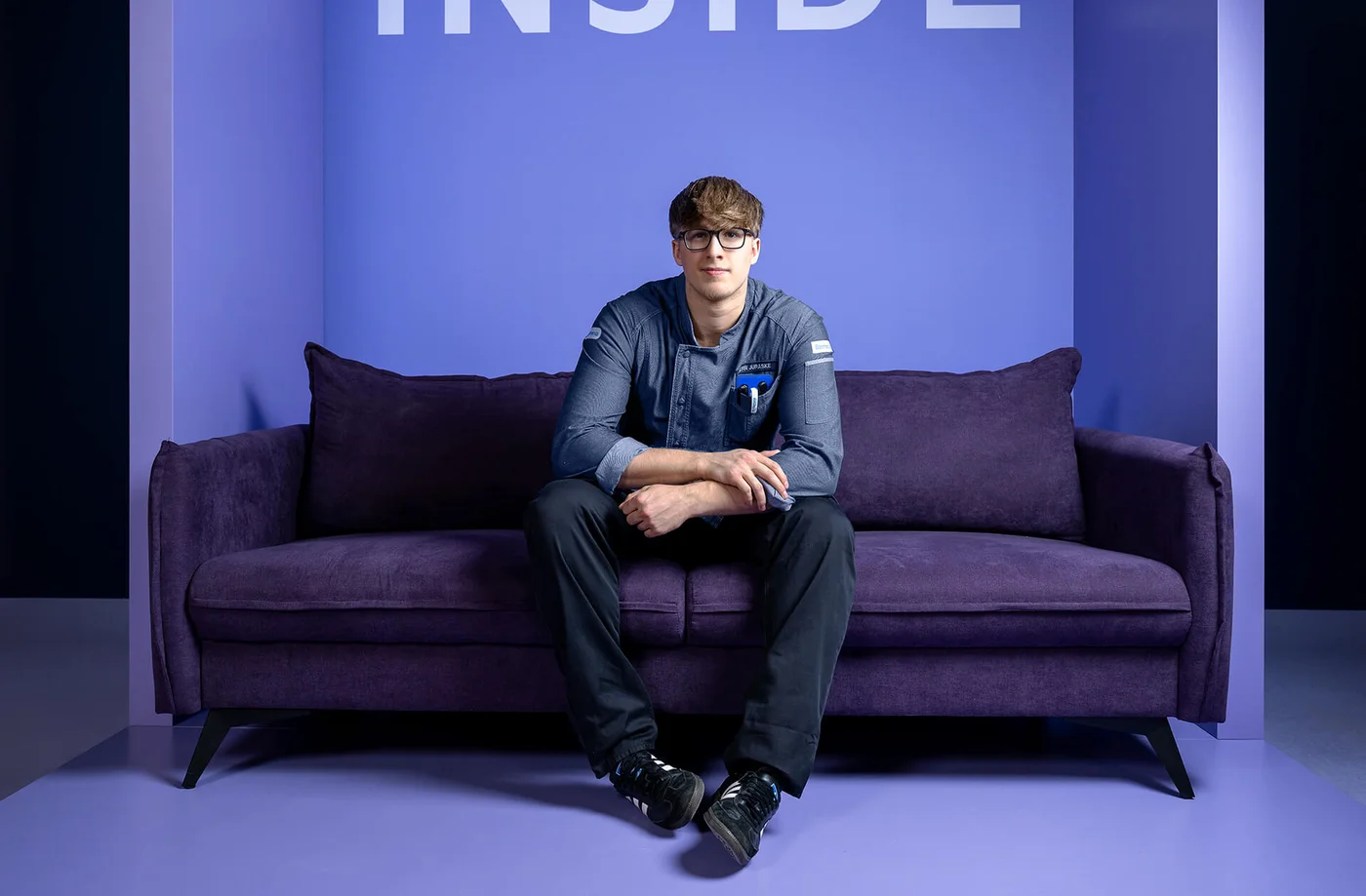 Eine Person sitzt auf einem lila Sofa vor einer lila Wand mit dem Wort "INSIDE" in großen Buchstaben darüber. Die Umgebung ist minimalistisch und in verschiedenen Lilatönen gehalten.