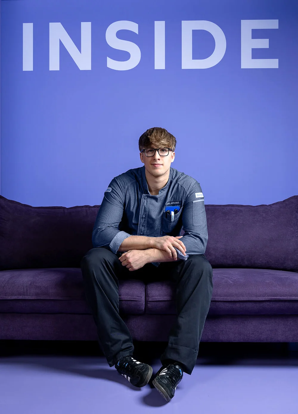 Eine Person sitzt auf einem lila Sofa vor einer lila Wand mit dem Wort "INSIDE" in großen Buchstaben darüber. Die Umgebung ist minimalistisch und in verschiedenen Lilatönen gehalten.