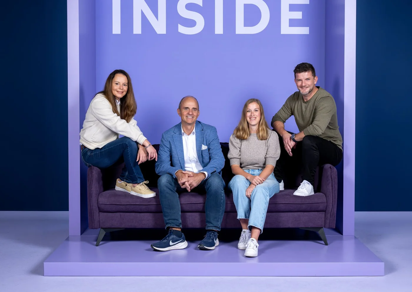 Vier Personen sitzen und stehen um ein lila Sofa vor einem lila Hintergrund. Eine Frau sitzt links, neben ihr ein Mann im Anzug. Eine weitere Frau sitzt neben ihm, und ein anderer Mann lehnt lächelnd rechts am Sofa. Alle wirken entspannt und freundlich.