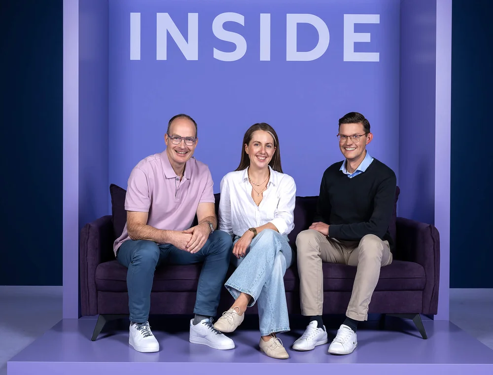 Drei Personen sitzen lächelnd auf einem lila Sofa. Hinter ihnen ist eine Wand mit dem Wort „INSIDE“ in großen Buchstaben zu sehen. Die Szene strahlt eine freundliche und entspannte Atmosphäre aus.
