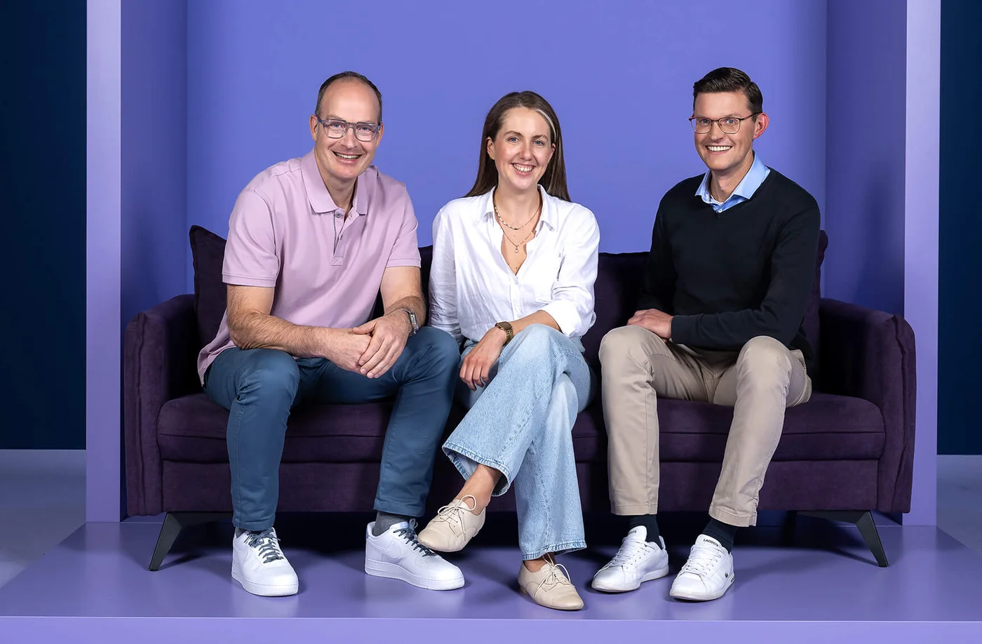 Drei Personen sitzen lächelnd auf einem lila Sofa. Hinter ihnen ist eine Wand mit dem Wort „INSIDE“ in großen Buchstaben zu sehen. Die Szene strahlt eine freundliche und entspannte Atmosphäre aus.