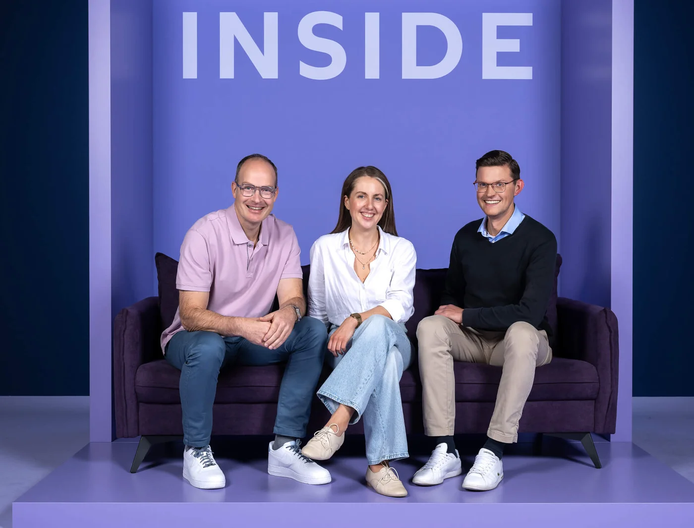 Drei Personen sitzen auf einem Sofa vor einem lila Hintergrund mit dem Wort "INSIDE" darüber. Alle lächeln und schauen in die Kamera. Die Gruppe vermittelt eine freundliche und entspannte Atmosphäre. Sie tragen legere Kleidung in hellen Tönen.