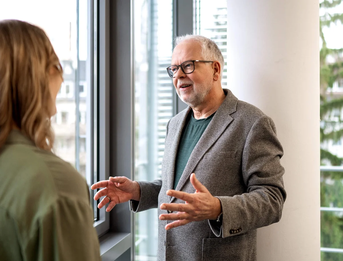 Ein älterer Mann mit Brille und grauem Sakko spricht gestikulierend mit einer Frau in einem Innenraum. Sie stehen in der Nähe eines großen Fensters. Die Szene wirkt wie ein kollegiales Gespräch oder eine Beratung in einem Büro.