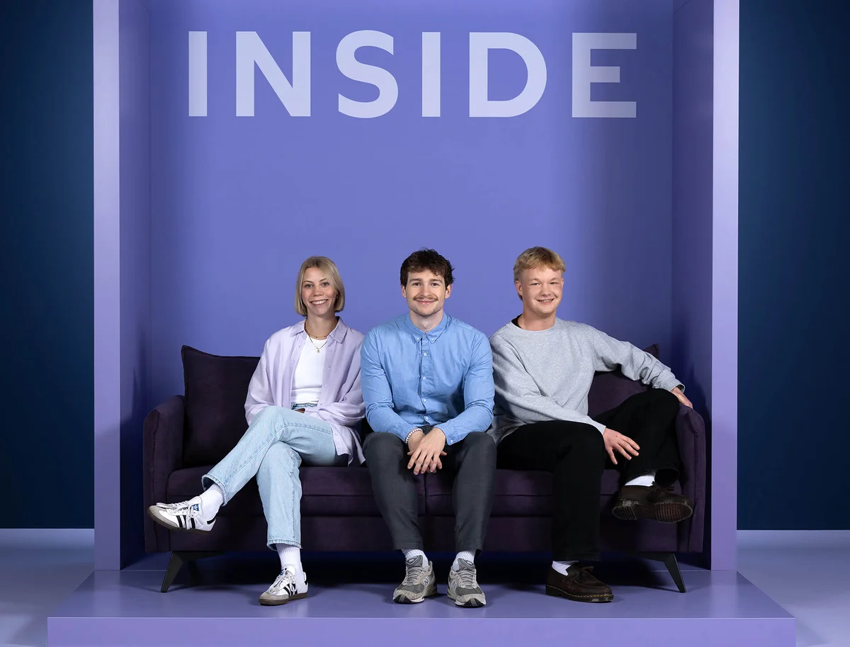 Drei Personen sitzen lächelnd auf einem lila Sofa vor einem Hintergrund mit dem Wort "INSIDE". Alle tragen Freizeitkleidung und wirken entspannt und zufrieden.