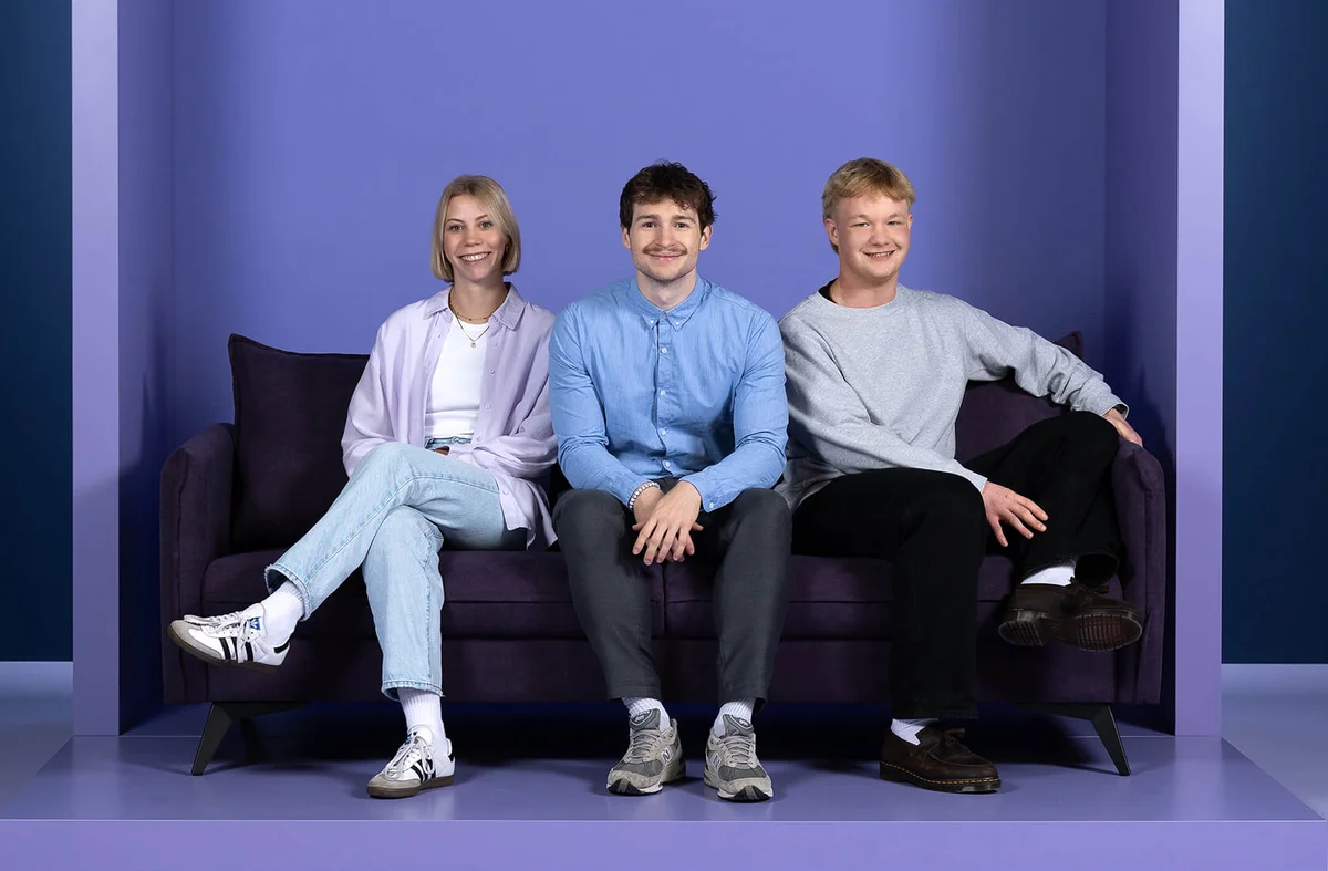 Drei Personen sitzen lächelnd auf einem lila Sofa vor einem Hintergrund mit dem Wort "INSIDE". Alle tragen Freizeitkleidung und wirken entspannt und zufrieden.