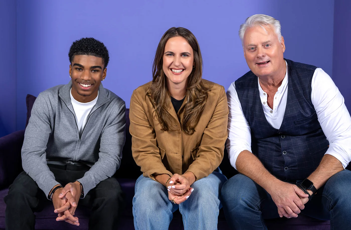 Drei Personen sitzen auf einem Sofa vor einem lilafarbenen Hintergrund. Sie lächeln in die Kamera. Die Person links trägt einen grauen Hoodie, die in der Mitte eine beige Jacke und die rechte Person ein weißes Hemd mit Weste.