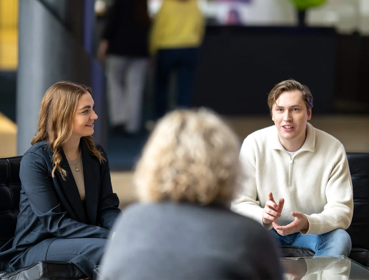 Drei Personen sitzen in einem Gespräch. Zwei von ihnen, ein Mann und eine Frau, lachen und unterhalten sich. Eine weitere Person sitzt ihnen gegenüber. Die Atmosphäre wirkt entspannt und freundlich.
