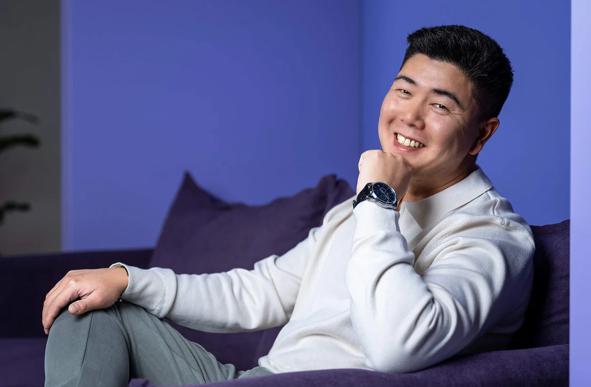 Eine lächelnde Person sitzt auf einem lila Sofa vor einem blauen Hintergrund. Sie trägt einen weißen Pullover und eine Armbanduhr, die mit der linken Hand abgestützt ist.