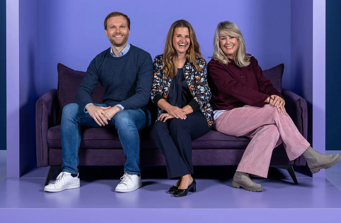 Drei Personen sitzen nebeneinander auf einem lila Sofa vor einem lila Hintergrund. Alle lächeln fröhlich. Eine Person trägt einen Pullover und Jeans, die zweite eine gemusterte Jacke, und die dritte einen roten Pullover mit hellen Hosen.