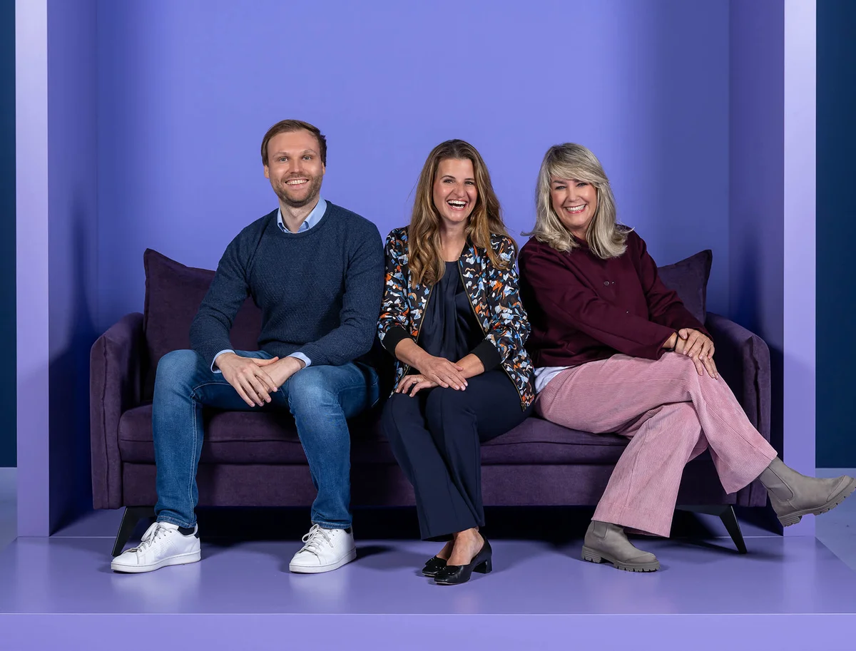 Drei Personen sitzen nebeneinander auf einem lila Sofa vor einem lila Hintergrund. Alle lächeln fröhlich. Eine Person trägt einen Pullover und Jeans, die zweite eine gemusterte Jacke, und die dritte einen roten Pullover mit hellen Hosen.
