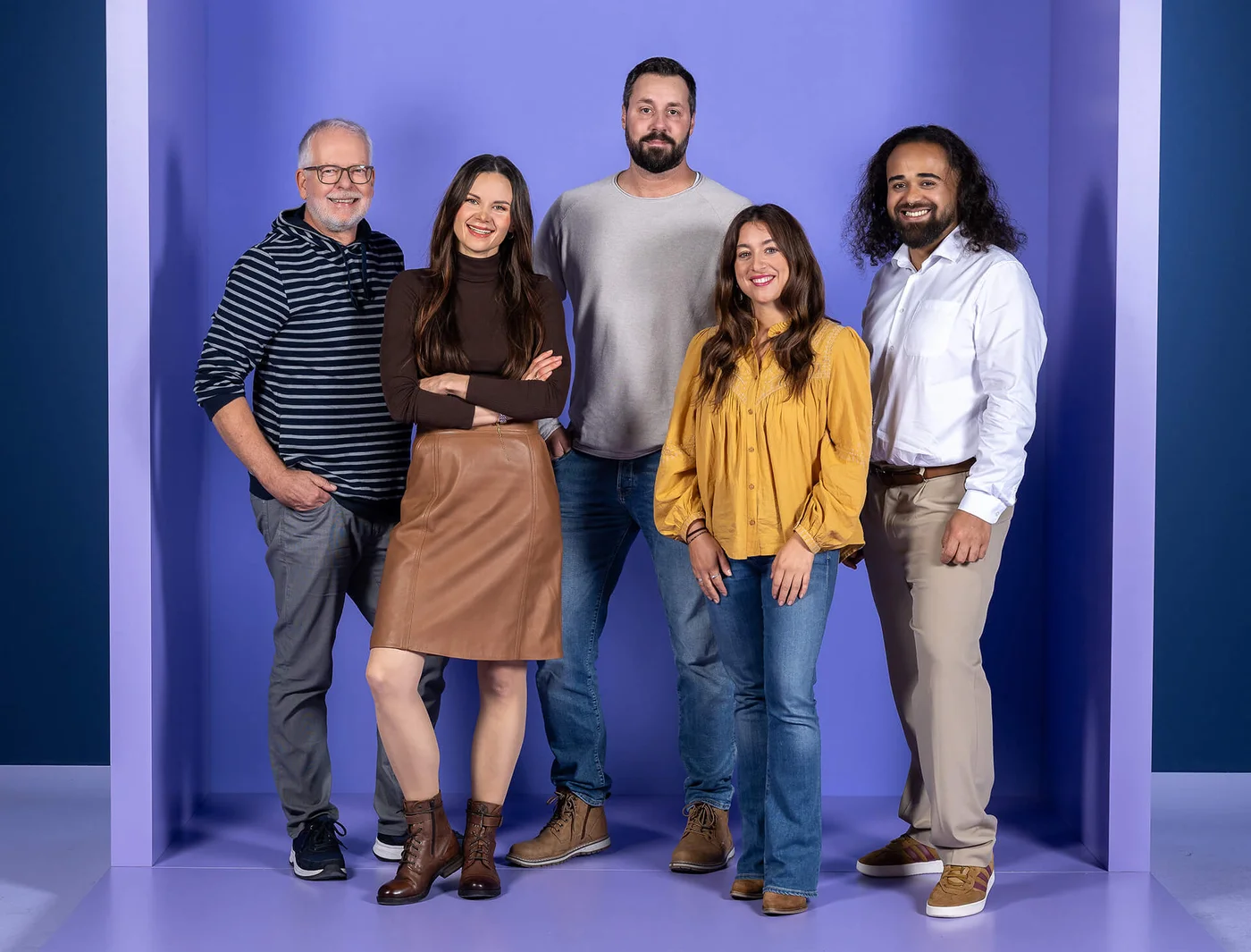 Fünf Personen stehen lächelnd vor einem lila Hintergrund. Drei Männer und zwei Frauen sind lässig gekleidet. Sie stehen entspannt nebeneinander und wirken freundlich.