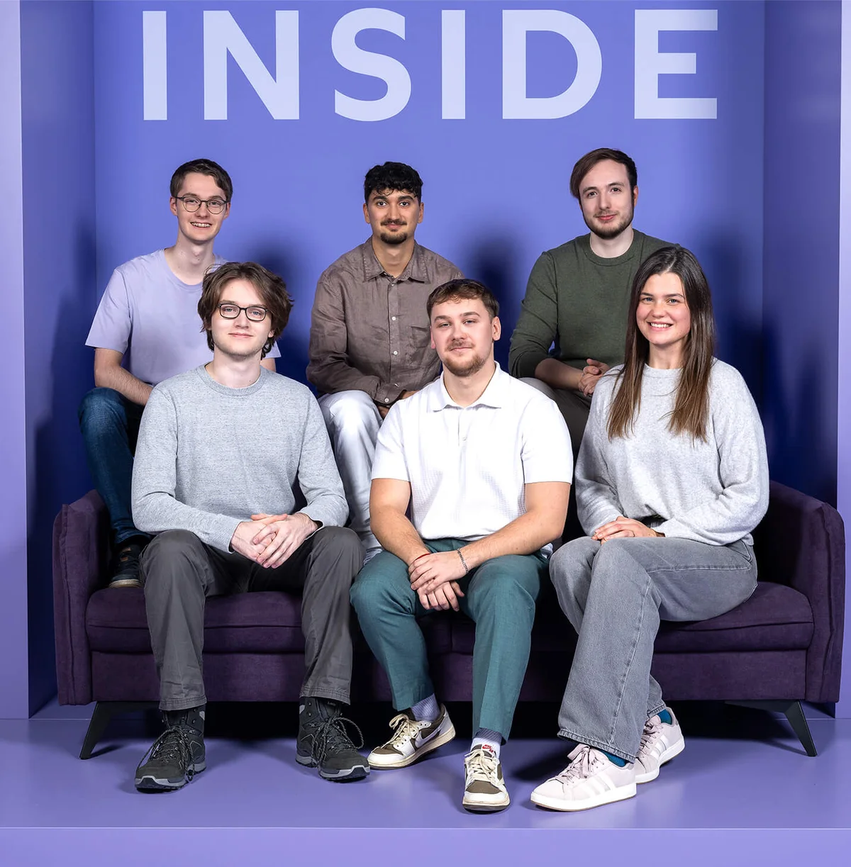 Sechs Personen sitzen und stehen vor einem lila Hintergrund mit dem Wort "INSIDE" oben. Sie sind entspannt gekleidet und blicken lächelnd in die Kamera.