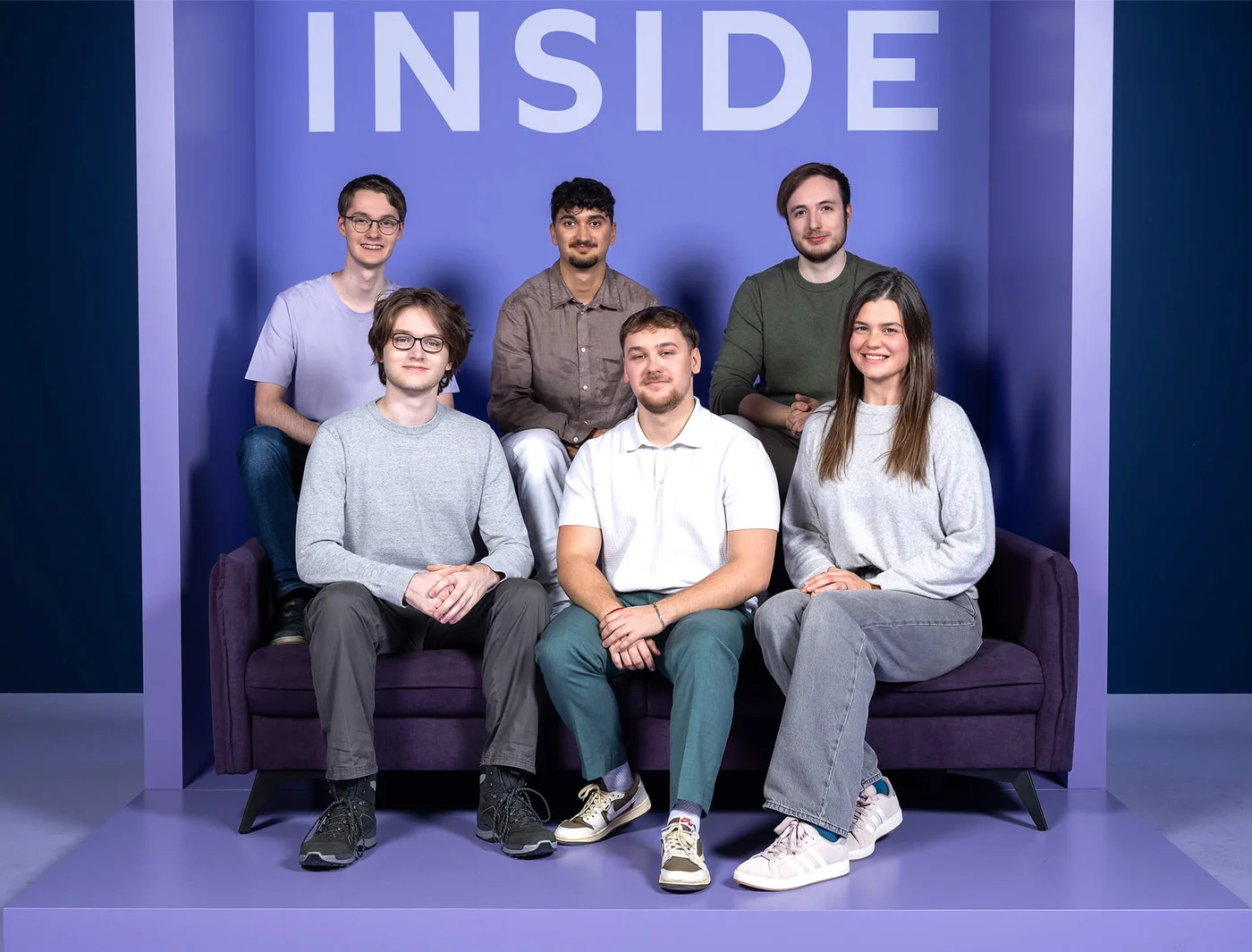 Sechs Personen sitzen und stehen vor einem lila Hintergrund mit dem Wort "INSIDE" oben. Sie sind entspannt gekleidet und blicken lächelnd in die Kamera.