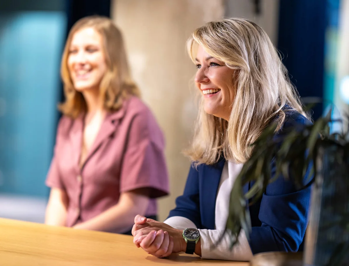 Zwei lächelnde Frauen sitzen an einem Tisch in einem gut beleuchteten Raum. Die Frau im Vordergrund trägt einen blauen Blazer und hat blondes Haar. Beide wirken entspannt und freundlich, als ob sie in einem Gespräch oder bei einem Meeting sind.