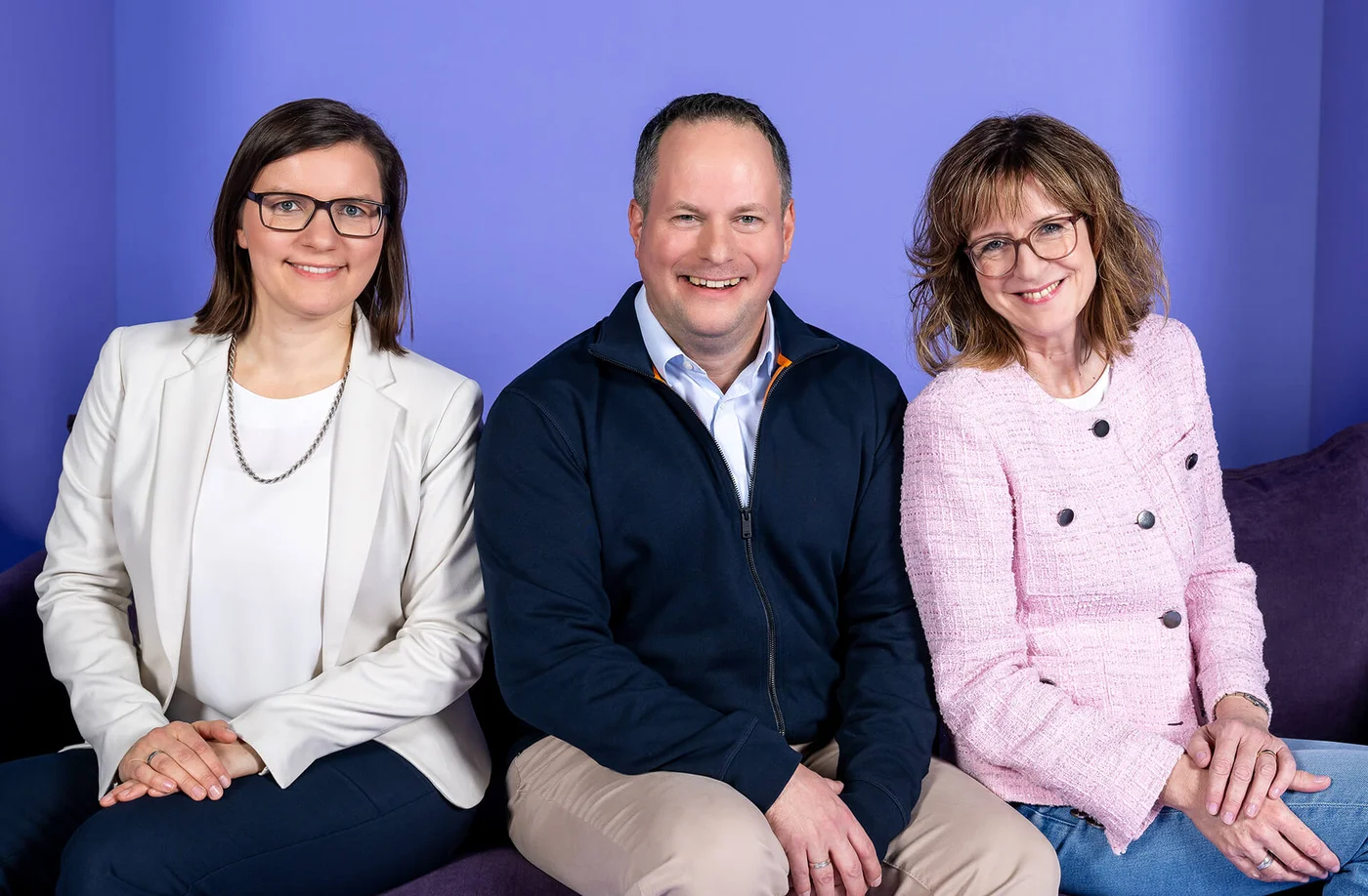 Drei Personen sitzen auf einem Sofa vor einem lilafarbenen Hintergrund. Eine Frau trägt einen weißen Blazer, der Mann in der Mitte trägt eine dunkle Jacke, und die Frau rechts trägt eine rosa Jacke. Alle lächeln in die Kamera.