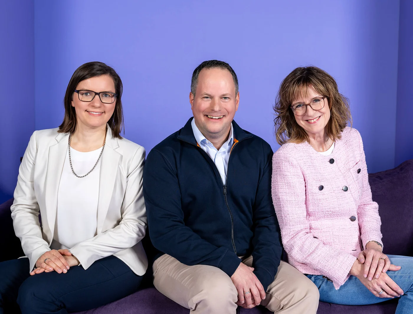 Drei Personen sitzen auf einem Sofa vor einem lilafarbenen Hintergrund. Eine Frau trägt einen weißen Blazer, der Mann in der Mitte trägt eine dunkle Jacke, und die Frau rechts trägt eine rosa Jacke. Alle lächeln in die Kamera.
