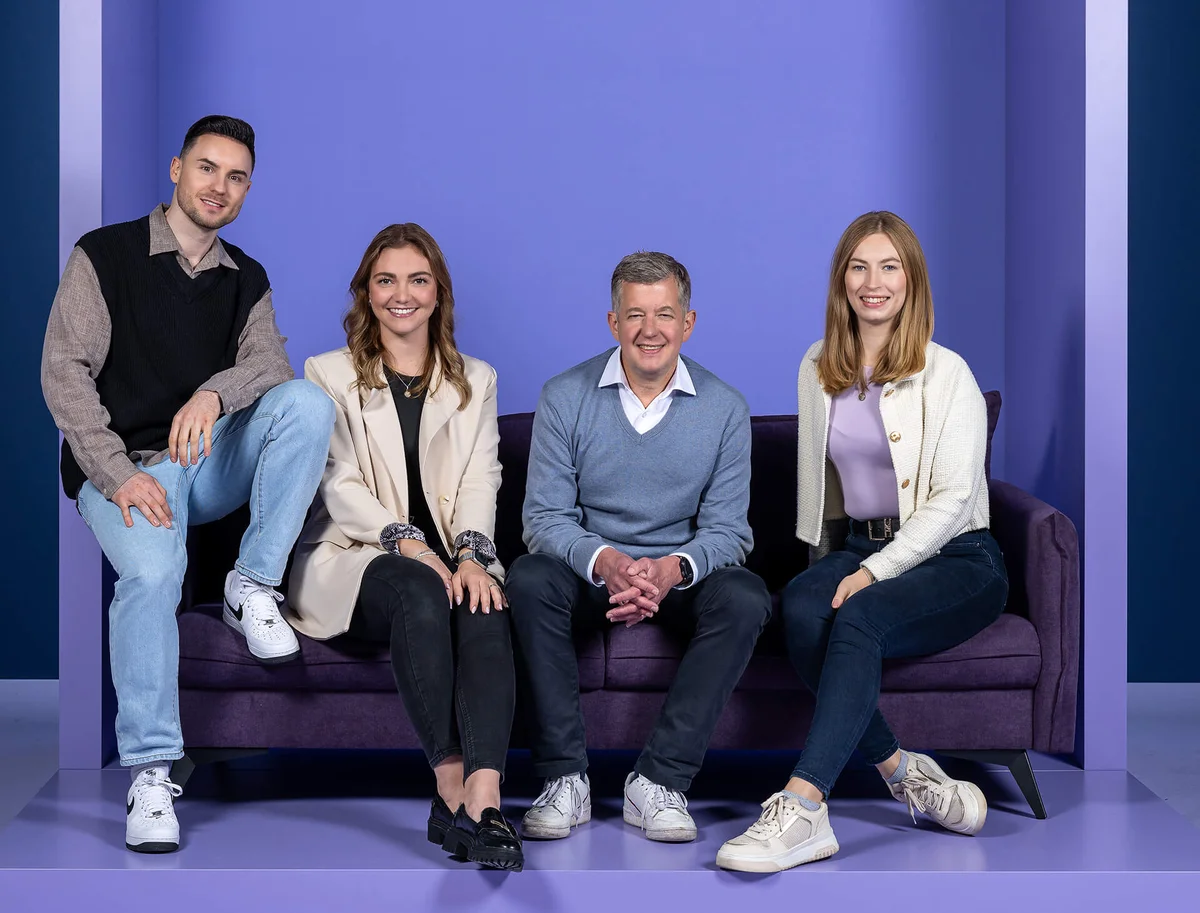 Eine Gruppe von vier Personen sitzt und steht um ein dunkellila Sofa vor einem einfarbigen lila Hintergrund. Alle lächeln in die Kamera. Die Gruppe besteht aus zwei Frauen und zwei Männern verschiedener Altersgruppen, die lässig, aber stilvoll gekleidet sind.