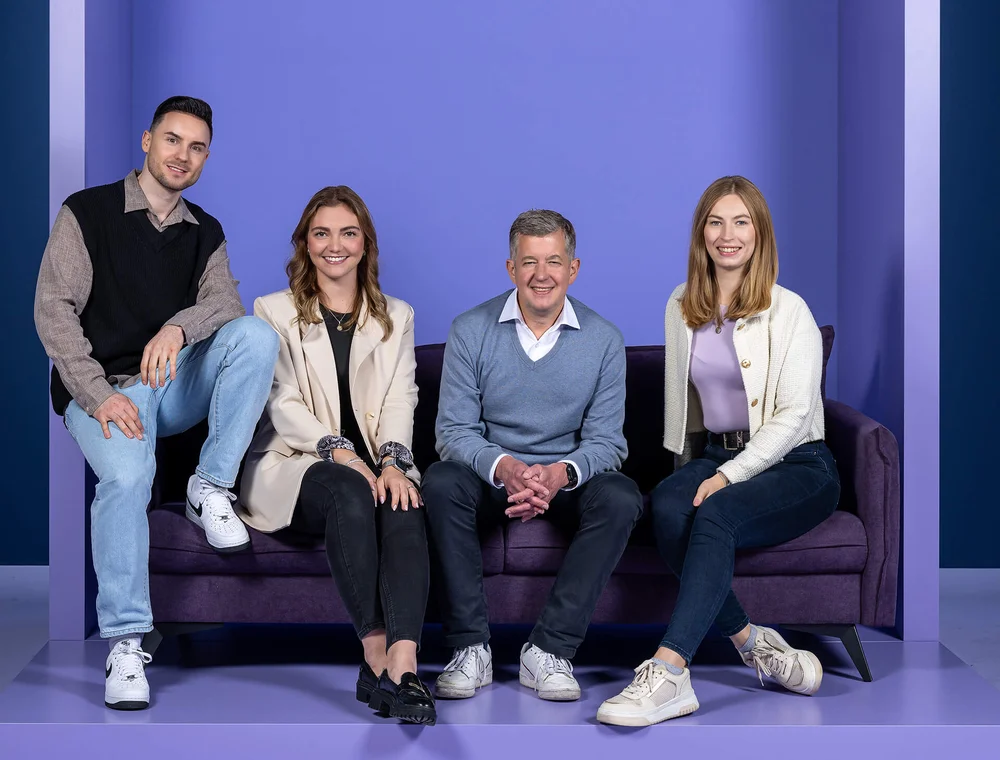 Eine Gruppe von vier Personen sitzt und steht um ein dunkellila Sofa vor einem einfarbigen lila Hintergrund. Alle lächeln in die Kamera. Die Gruppe besteht aus zwei Frauen und zwei Männern verschiedener Altersgruppen, die lässig, aber stilvoll gekleidet sind.