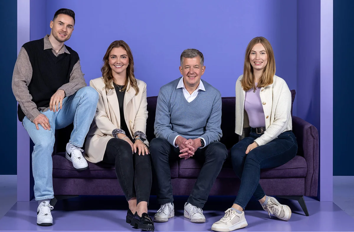 Eine Gruppe von vier Personen sitzt und steht um ein dunkellila Sofa vor einem einfarbigen lila Hintergrund. Alle lächeln in die Kamera. Die Gruppe besteht aus zwei Frauen und zwei Männern verschiedener Altersgruppen, die lässig, aber stilvoll gekleidet sind.