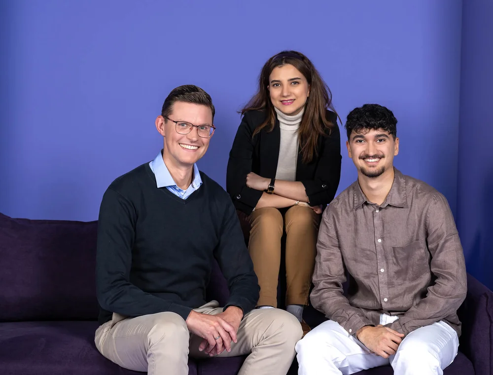 Drei lächelnde Personen sitzen vor einem blauen Hintergrund auf einem Sofa. Links ein Mann mit Brille in einem dunkelblauen Pulli, in der Mitte eine Frau in heller Kleidung, rechts ein Mann in einem beigefarbenen Hemd. Alle wirken freundlich und entspannt.