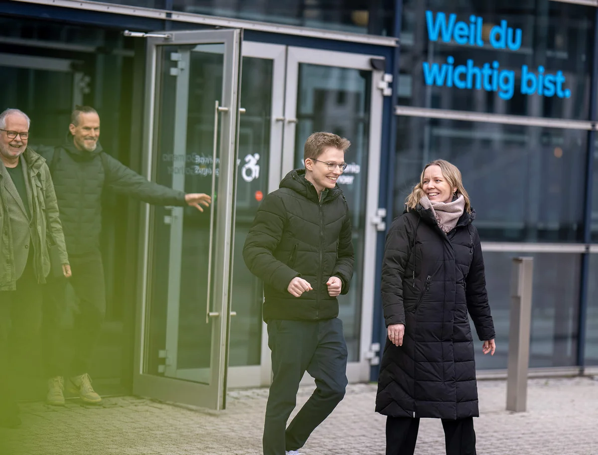 Eine Gruppe von Menschen in Wintermänteln verlässt ein modernes Gebäude. Zwei Personen vorne laufen und lächeln, während zwei Personen im Hintergrund durch die Tür gehen. Auf der Glaswand steht der Schriftzug: „Weil du wichtig bist.“
