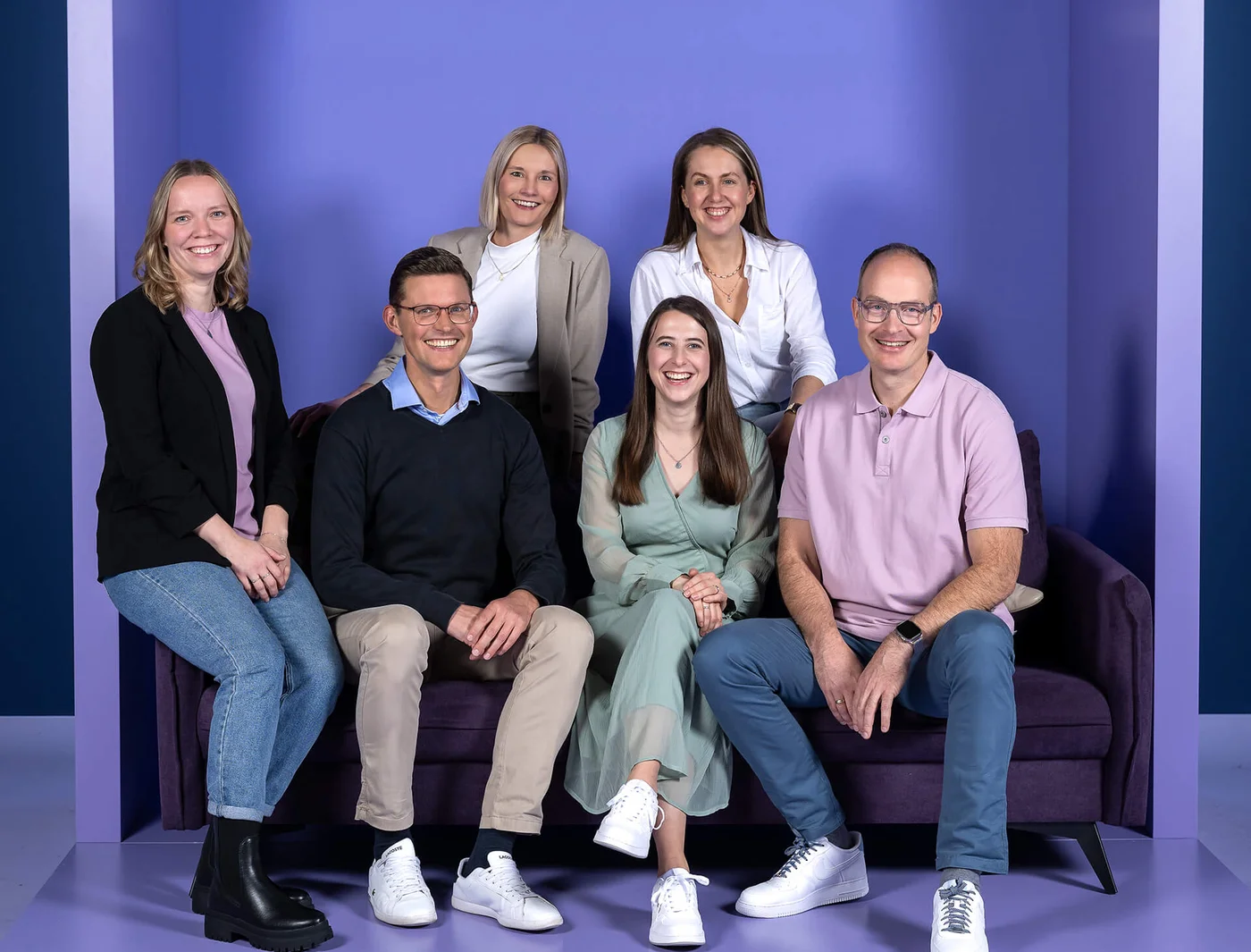 Eine Gruppe von sechs Personen sitzt und steht um ein lila Sofa. Sie lächeln und tragen legere Kleidung in neutralen Farben. Der Hintergrund ist ebenfalls lila.