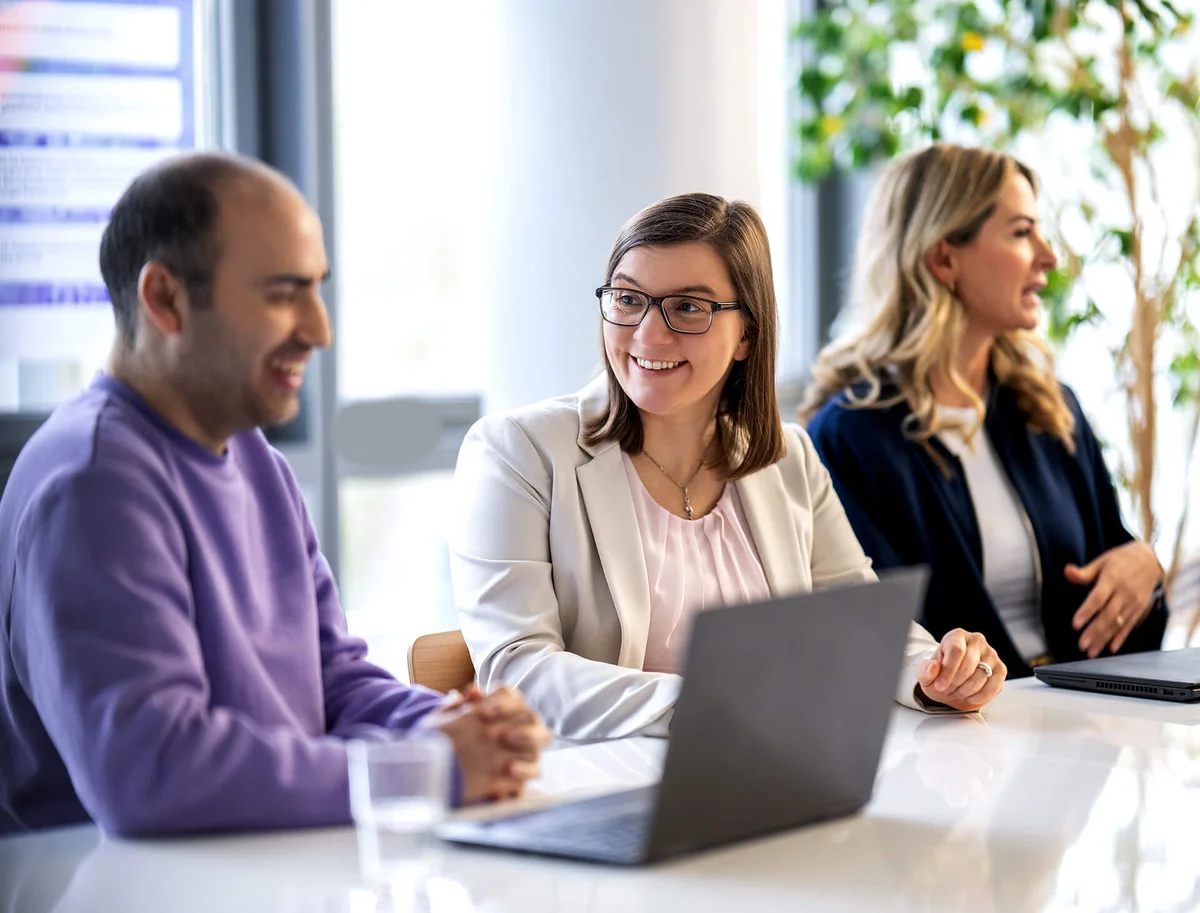 Drei Personen sitzen lächelnd an einem Tisch in einem hellen Büro. Zwei Frauen und ein Mann blicken sich bei einer Besprechung an. Alle haben Laptops vor sich, was auf ein Arbeitsmeeting hindeutet. Die Stimmung ist freundlich und entspannt.