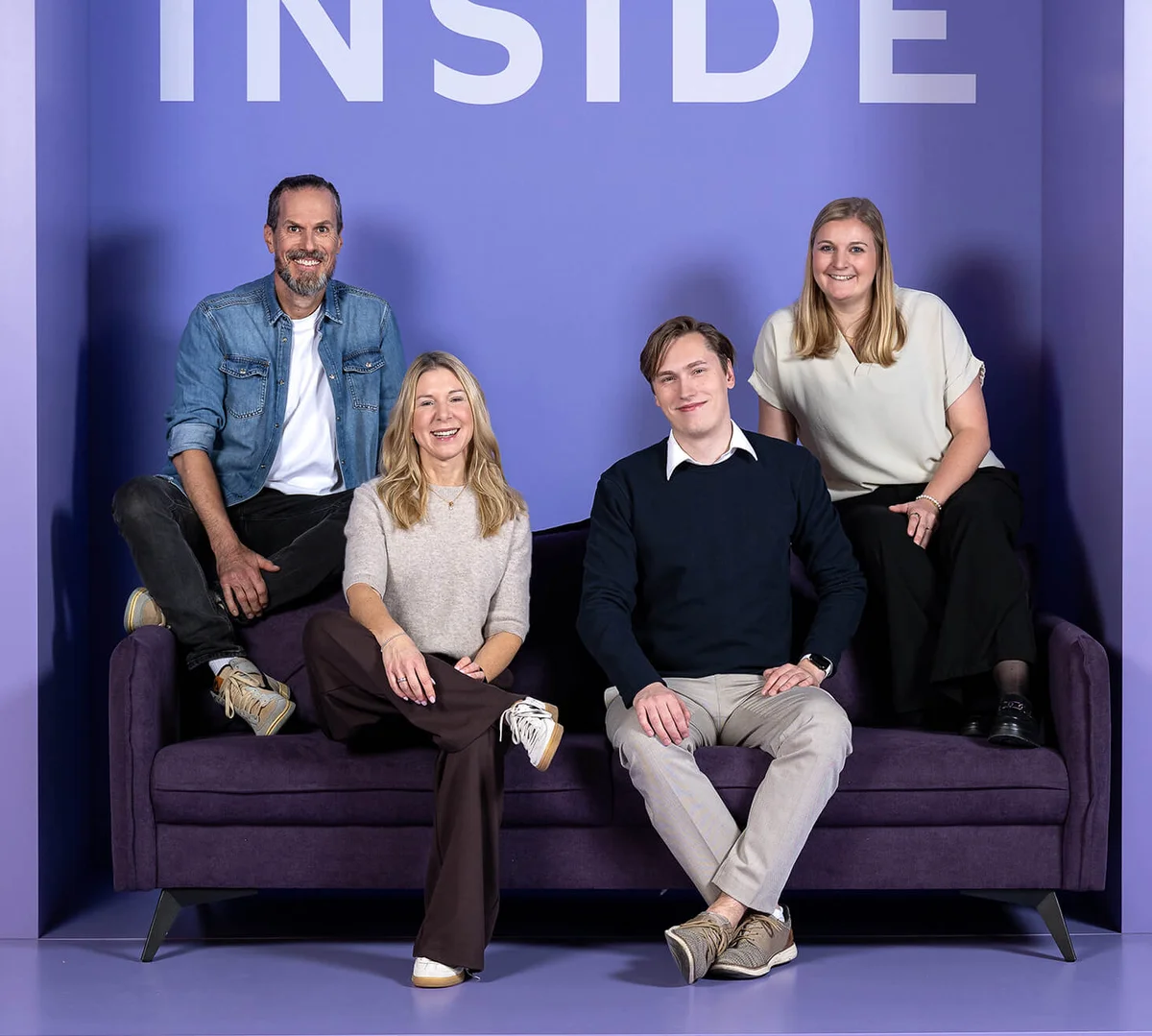 Eine Gruppe von vier Personen sitzt und steht auf einem violetten Sofa vor einem hellvioletten Hintergrund mit dem Schriftzug "INSIDE". Alle lächeln freundlich in die Kamera.
