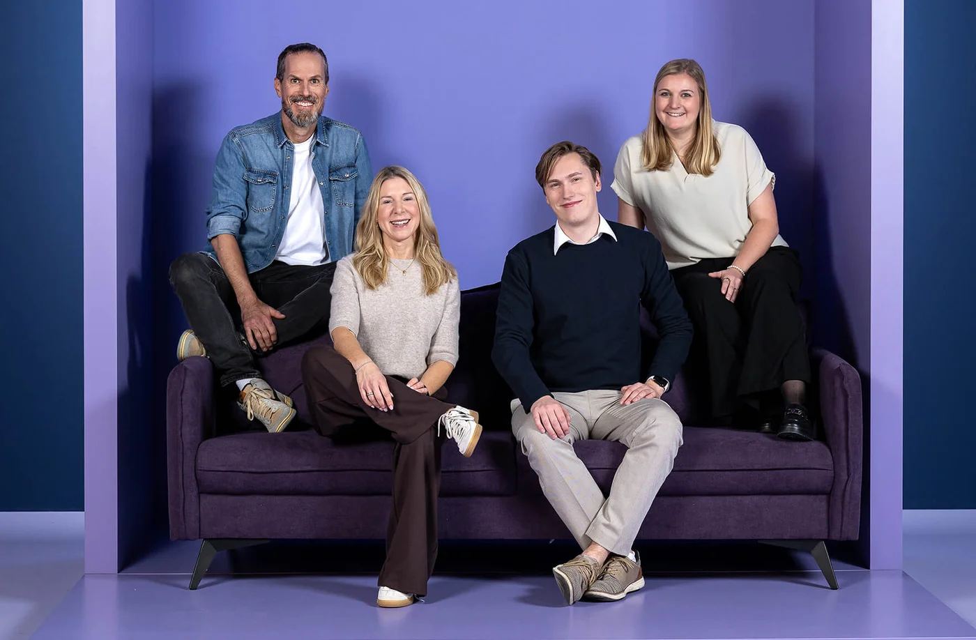 Eine Gruppe von vier Personen sitzt und steht auf einem violetten Sofa vor einem hellvioletten Hintergrund mit dem Schriftzug "INSIDE". Alle lächeln freundlich in die Kamera.