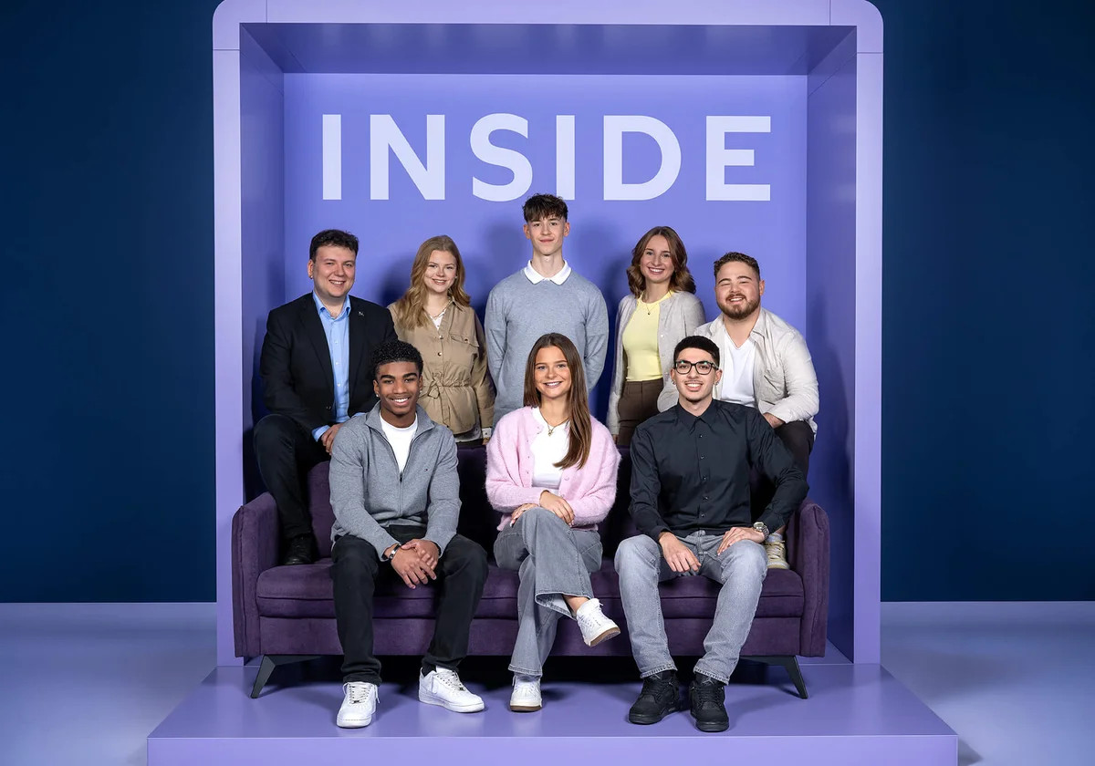 Eine Gruppe von acht Personen sitzt und steht vor einer Wand mit dem Wort „INSIDE“ auf einem lilafarbenen Hintergrund. Die Personen lächeln in die Kamera und sind in lässiger Kleidung.