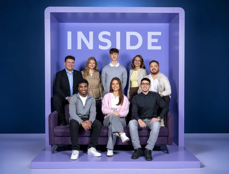 Eine Gruppe von acht Personen sitzt und steht vor einer Wand mit dem Wort „INSIDE“ auf einem lilafarbenen Hintergrund. Die Personen lächeln in die Kamera und sind in lässiger Kleidung.