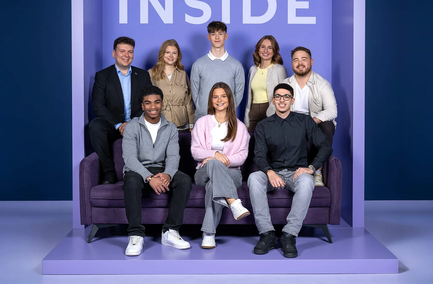 Eine Gruppe von acht Personen sitzt und steht vor einer Wand mit dem Wort „INSIDE“ auf einem lilafarbenen Hintergrund. Die Personen lächeln in die Kamera und sind in lässiger Kleidung.