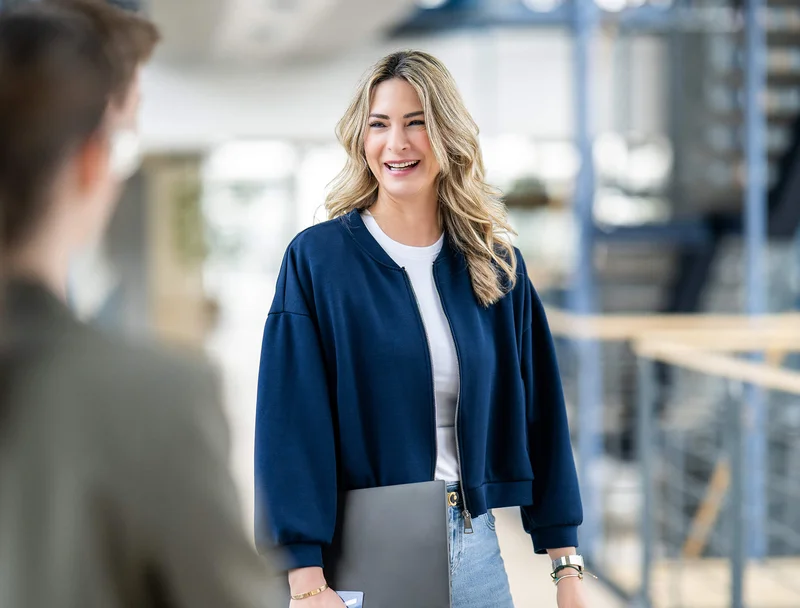 Eine junge Frau mit blonden Haaren lacht und hält einen Laptop. Sie trägt eine blaue Jacke und steht in einem modernen Bürogebäude. Im Vordergrund sieht man unscharf eine andere Person, mit der sie sich offenbar unterhält.