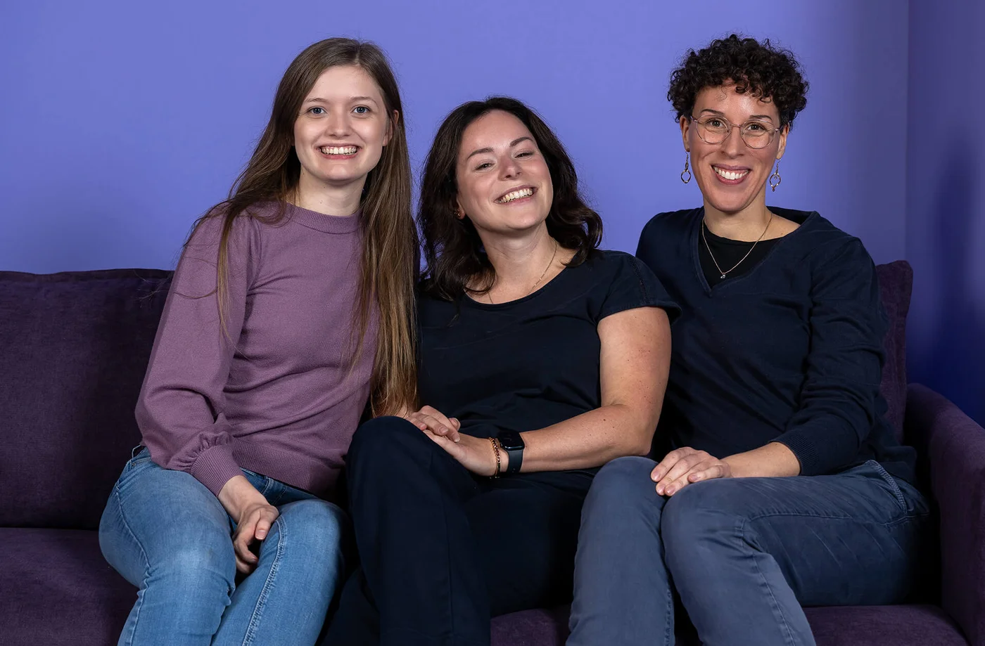 Drei Personen sitzen lächelnd nebeneinander auf einem Sofa. Sie wirken entspannt und fröhlich. Der Hintergrund ist in einem Lilaton gehalten.