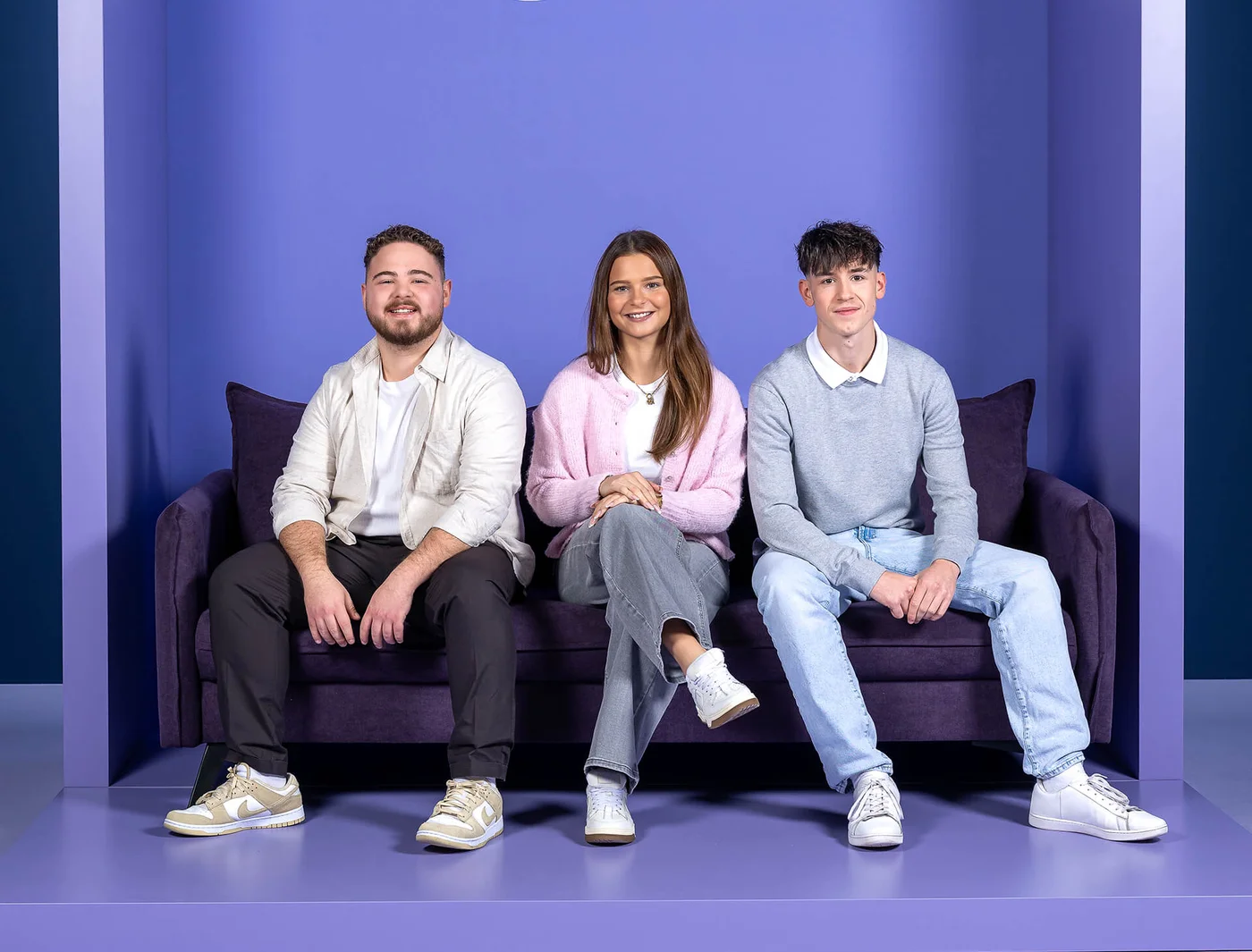 Drei junge Erwachsene sitzen auf einem lila Sofa vor einem lila Hintergrund. Die Frau in der Mitte trägt eine rosafarbene Jacke, während die beiden Männer links und rechts helle Hemden und Jeans tragen. Alle lächeln in die Kamera.