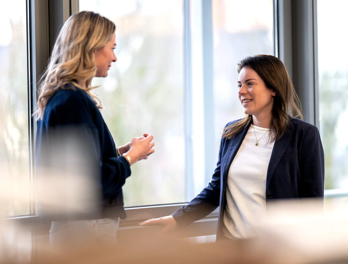 Zwei Frauen stehen in einem Büro am Fenster und unterhalten sich lächelnd. Die Frau links hat langes blondes Haar und trägt einen dunklen Pulli. Die Frau rechts hat schulterlanges dunkelbraunes Haar und trägt ein weißes Oberteil unter einem dunklen Blazer.